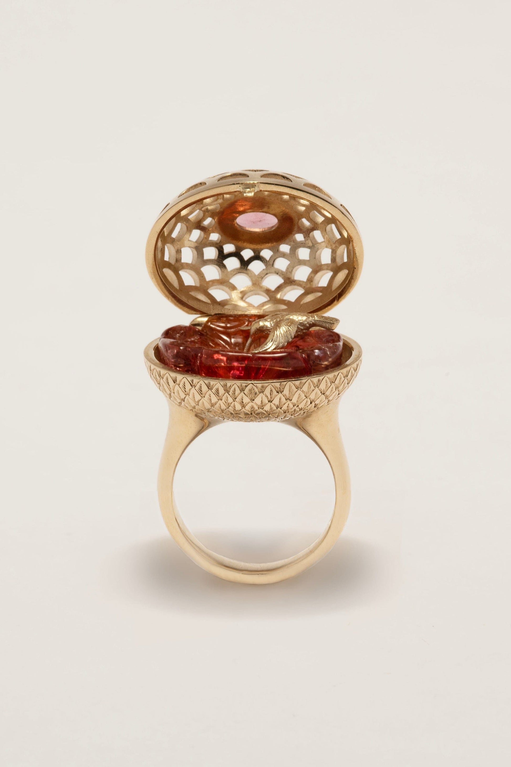 Gold ring with a hinged lid containing a red gemstone on a beige background
