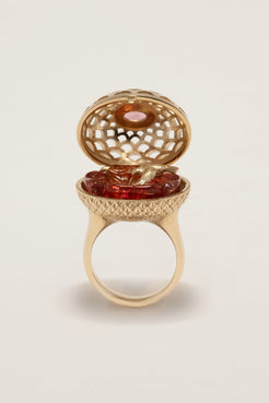 Gold ring with a hinged lid containing a red gemstone on a beige background