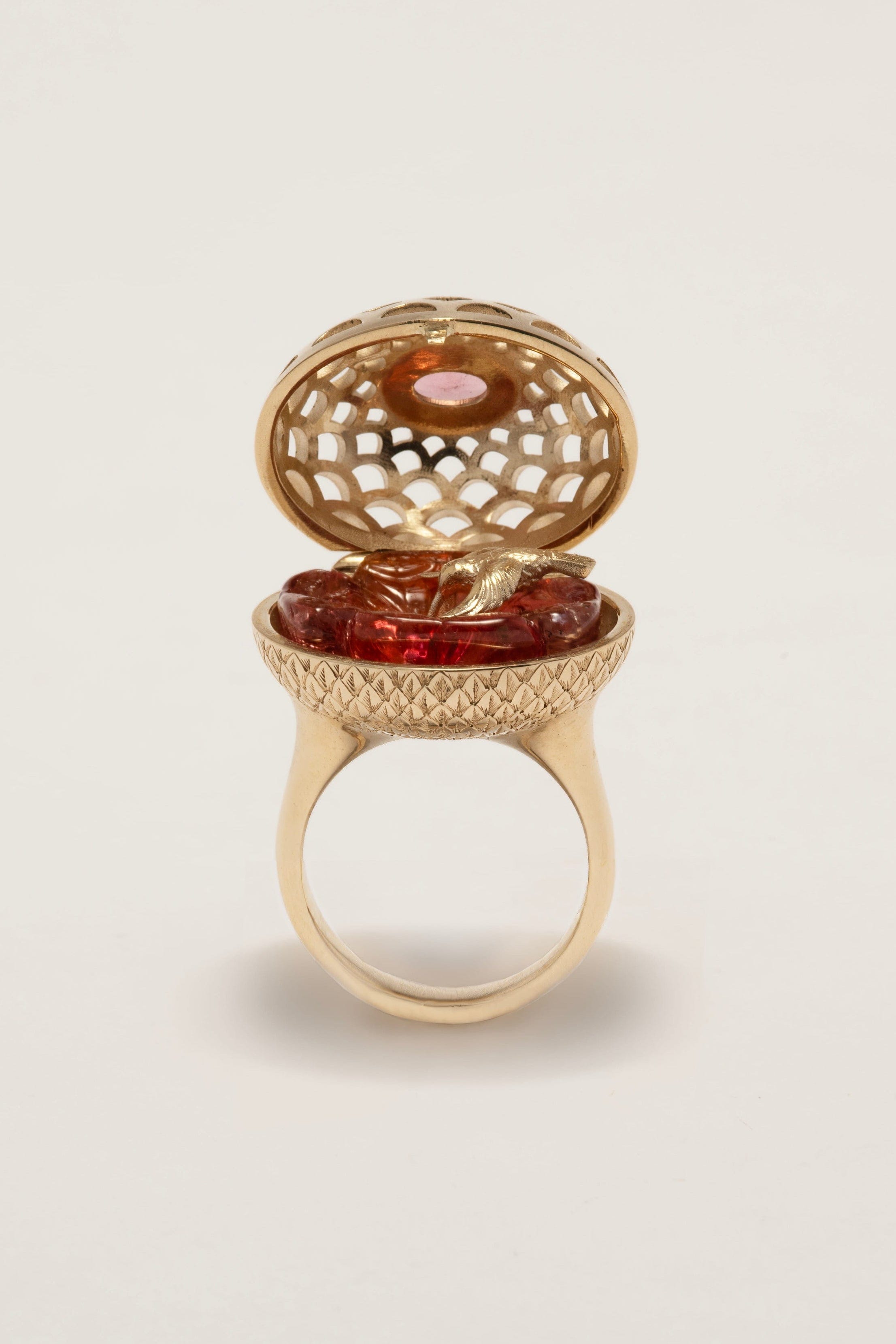 Gold ring with a hinged lid containing a red gemstone on a beige background